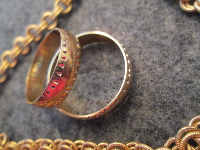 May include: Two gold-colored rings with a textured design. The ring on the left has a more intricate design with small triangles. The ring on the right has a smooth, rounded design.