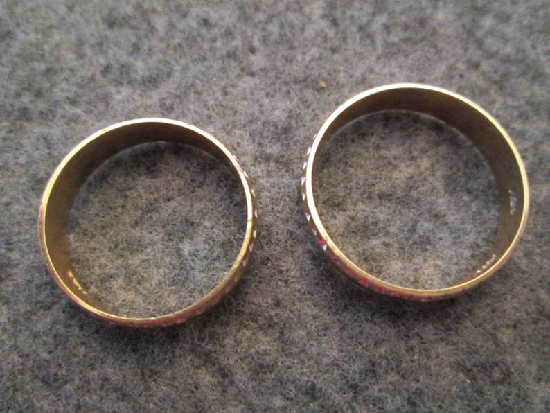 May include: Two gold-colored wedding bands with a textured pattern. The bands are simple and classic.