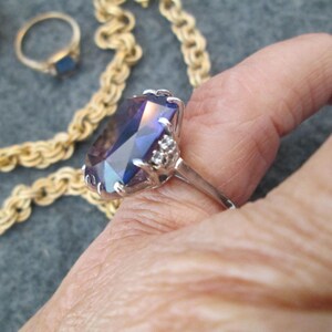 May include: A silver ring featuring a large, faceted, purple gemstone surrounded by smaller, clear gemstones. The ring is worn on a finger.