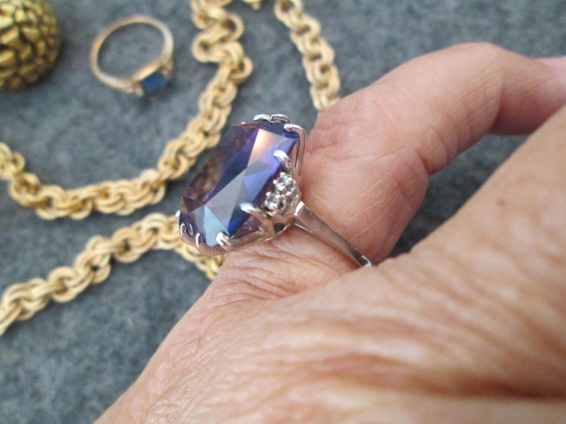 May include: A silver ring featuring a large, faceted, purple gemstone surrounded by smaller, clear gemstones. The ring is worn on a finger.
