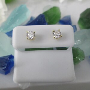 May include: A pair of stud earrings with round, clear gemstones set in gold-colored metal. The earrings are displayed on a white stand, surrounded by colorful glass pieces in shades of blue, green, and white.