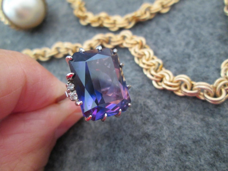 May include: A ring featuring a large, purple, emerald-cut gemstone surrounded by smaller, clear gemstones. The ring is set in silver.
