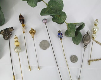May include: A set of eight vintage hat pins with various decorative tops, including a butterfly, a swirl of beads, a small dome, a single bead, a floral bead, a small dome, a silver dome, and a pearl and chain design. The pins are all gold-toned and have a long, thin shaft.