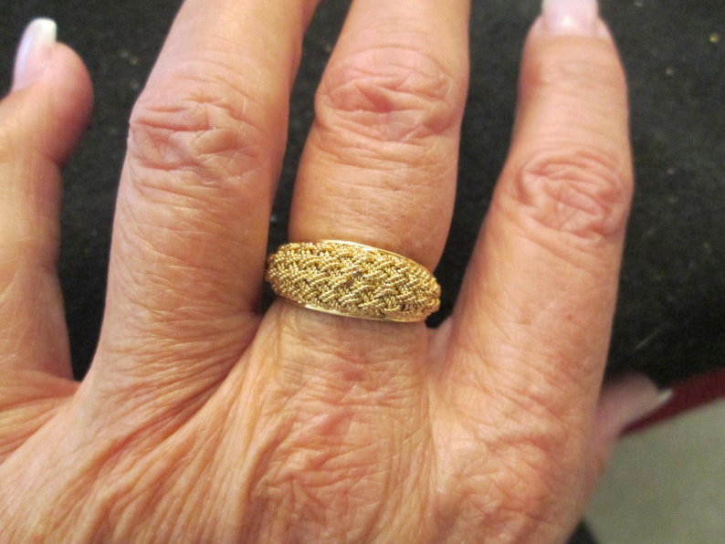 May include: A gold ring featuring a woven design. The ring is worn on a finger, with the woven pattern covering the top. The ring appears to be made of metal and has a shiny finish. The ring is a piece of jewellery.