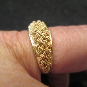 May include: A gold ring featuring a woven rope design. The ring, displayed on a finger, highlights the intricate texture and gleam of the metal. The design comprises tightly woven strands, creating a textured surface, showcasing the craftsmanship.