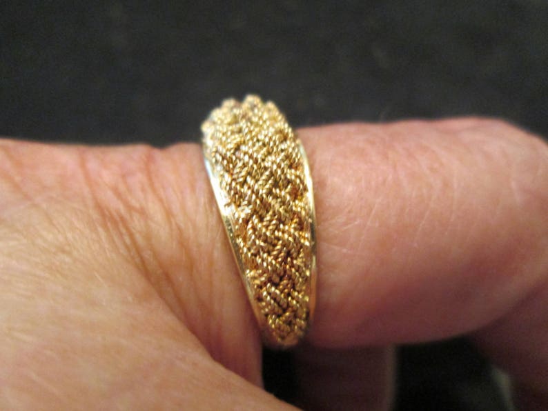 May include: A gold ring featuring a woven rope design. The ring, displayed on a finger, highlights the intricate texture and gleam of the metal. The design comprises tightly woven strands, creating a textured surface, showcasing the craftsmanship.