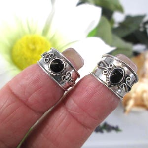 May include: Two silver rings featuring a wide band design with a central oval black stone. Each ring is accented with decorative twisted wire detailing and scrollwork patterns on the sides, creating a textured appearance on the polished metal surface.