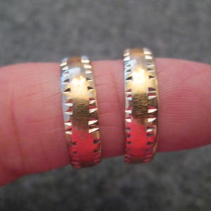 May include: Two gold wedding bands with a textured design. The bands are worn on a finger.
