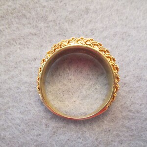 May include: A gold ring featuring a detailed, woven pattern around the band. The interior of the ring is smooth and polished, reflecting light. This ring is a piece of jewellery.