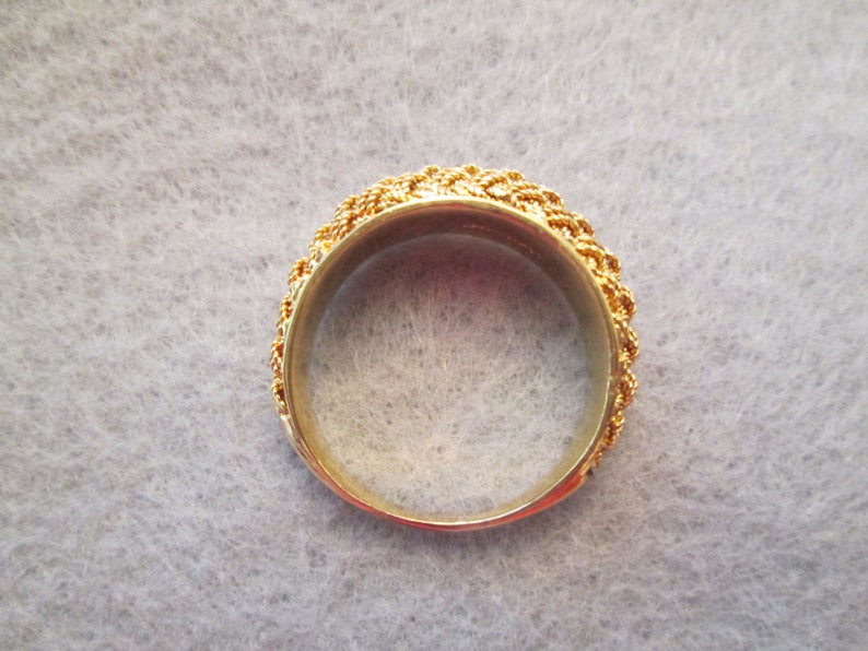 May include: A gold ring featuring a detailed, woven pattern around the band. The interior of the ring is smooth and polished, reflecting light. This ring is a piece of jewellery.