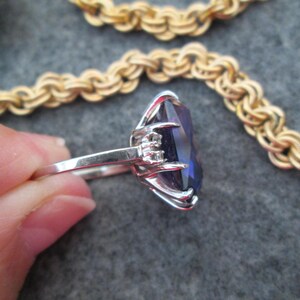 May include: A silver ring featuring a large, dark purple gemstone set in a prong setting. The ring is worn on a finger, with the gemstone facing the viewer.