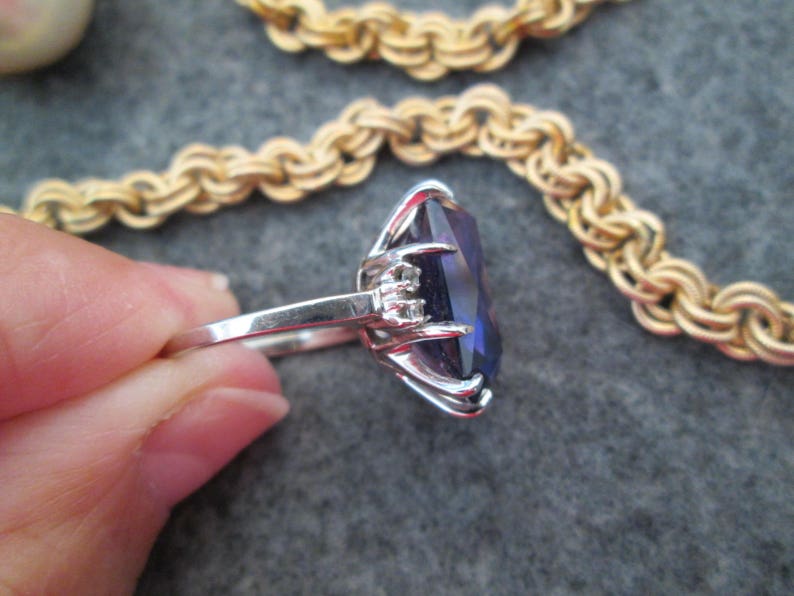 May include: A silver ring featuring a large, dark purple gemstone set in a prong setting. The ring is worn on a finger, with the gemstone facing the viewer.