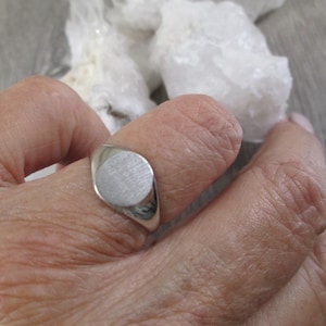 May include: A silver signet ring with a smooth, oval-shaped top. The ring is worn on a finger.