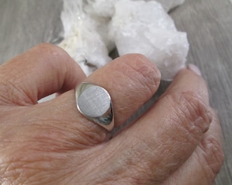 May include: A silver signet ring with a smooth, oval-shaped top. The ring is worn on a finger.