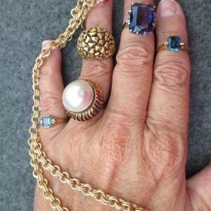 May include: A hand wearing five gold rings with different gemstones. The rings feature a pearl, a blue stone, a purple stone, and two textured gold bands. A gold chain necklace is wrapped around the wrist.