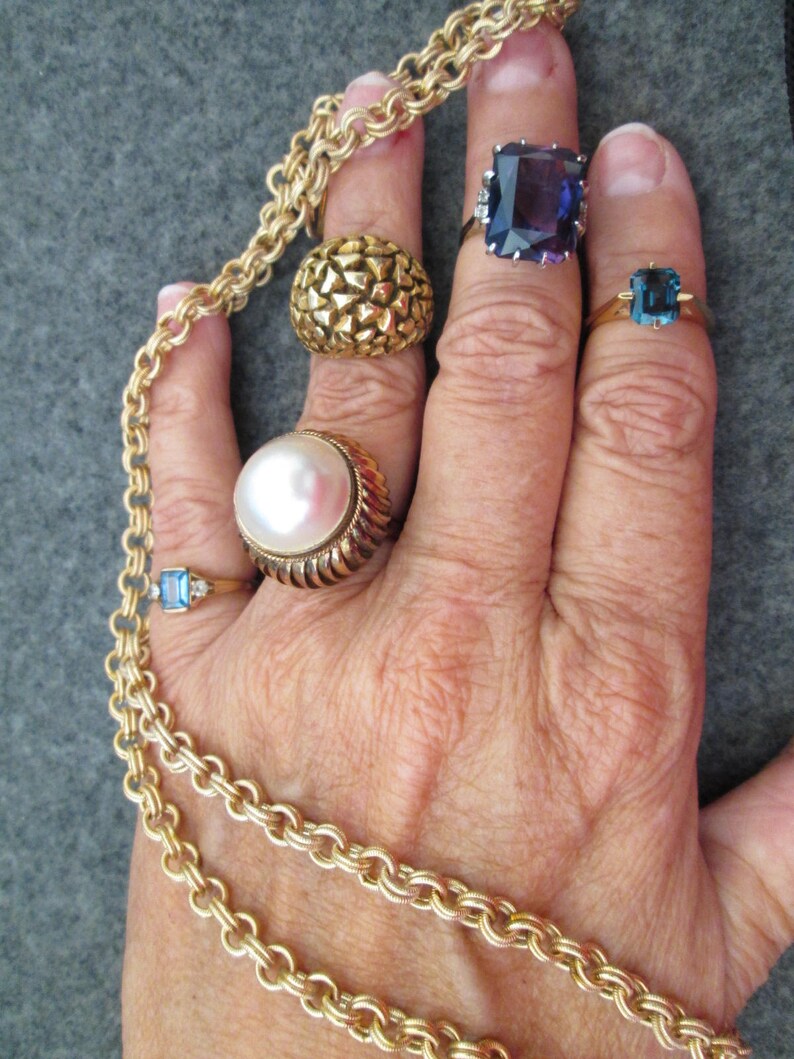 May include: A hand wearing five gold rings with different gemstones. The rings feature a pearl, a blue stone, a purple stone, and two textured gold bands. A gold chain necklace is wrapped around the wrist.
