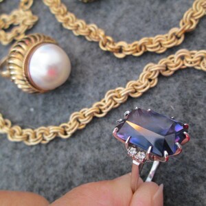 May include: A gold chain necklace with a pearl pendant and a ring featuring a large, faceted, purple gemstone with small diamond accents.