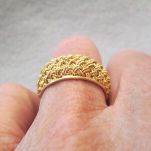May include: A gold ring with a woven texture. The ring is worn on a finger and has a band of woven gold. The ring's design is intricate and detailed, with a textured surface that catches the light.