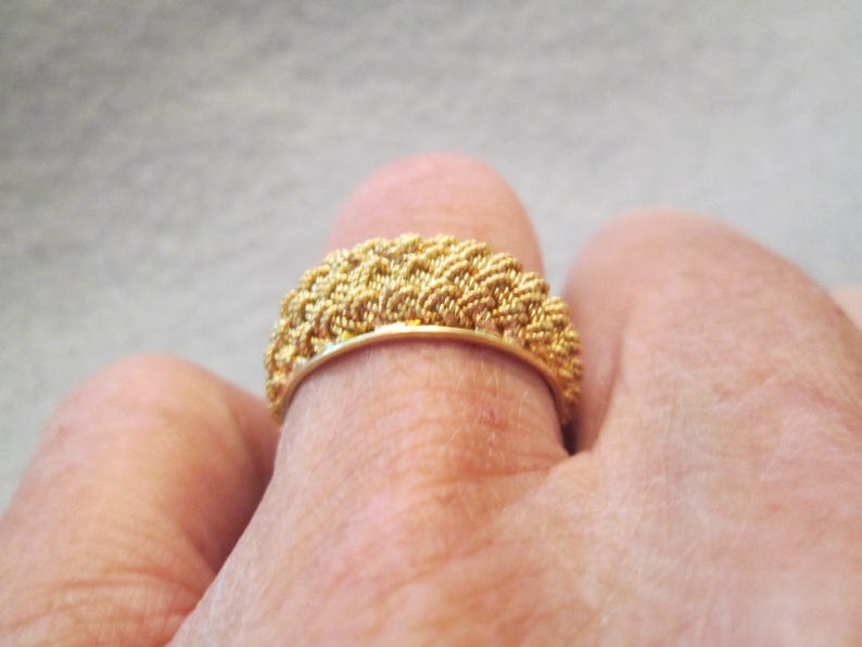 May include: A gold ring with a woven texture. The ring is worn on a finger and has a band of woven gold. The ring's design is intricate and detailed, with a textured surface that catches the light.