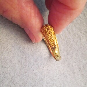 May include: A gold ring with a woven pattern on the top section. The ring is held between two fingers, highlighting its detailed texture and polished finish. The ring's band is smooth and shiny.