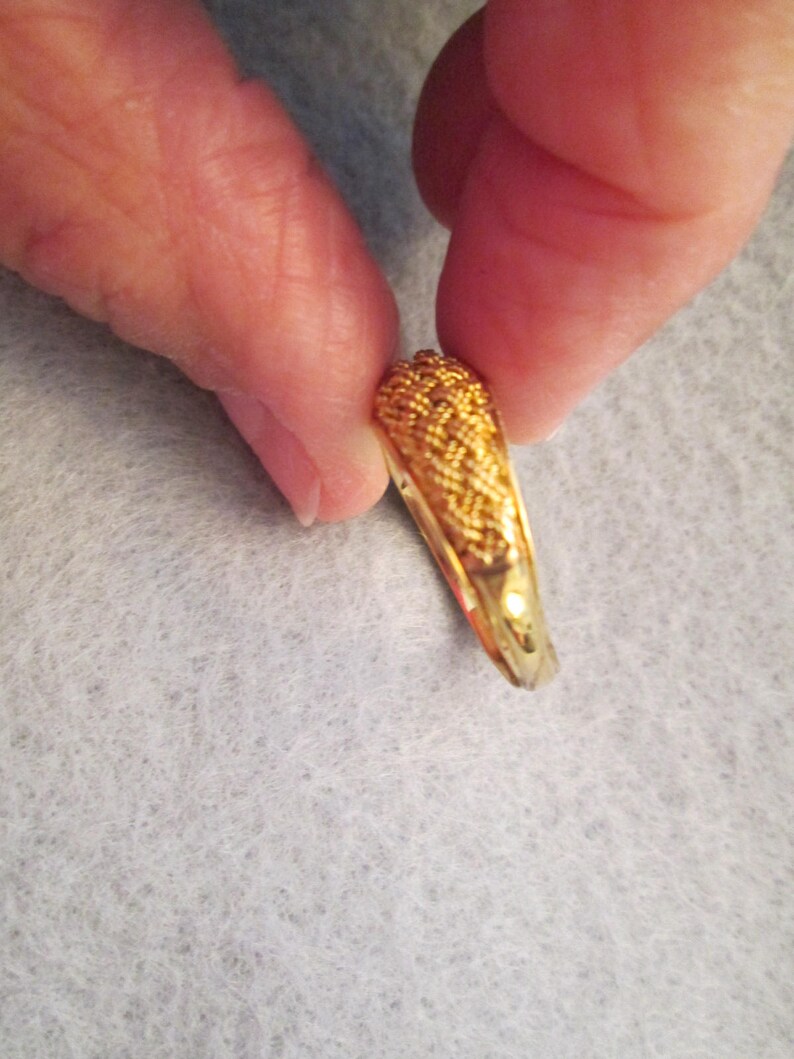 May include: A gold ring with a woven pattern on the top section. The ring is held between two fingers, highlighting its detailed texture and polished finish. The ring's band is smooth and shiny.