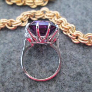 May include: A silver ring featuring a large, faceted, purple gemstone surrounded by smaller, clear gemstones.