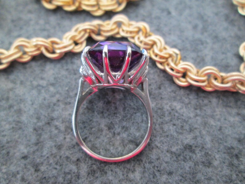 May include: A silver ring featuring a large, faceted, purple gemstone surrounded by smaller, clear gemstones.