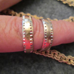 May include: Two gold wedding bands with a textured design. The bands are worn on a finger.