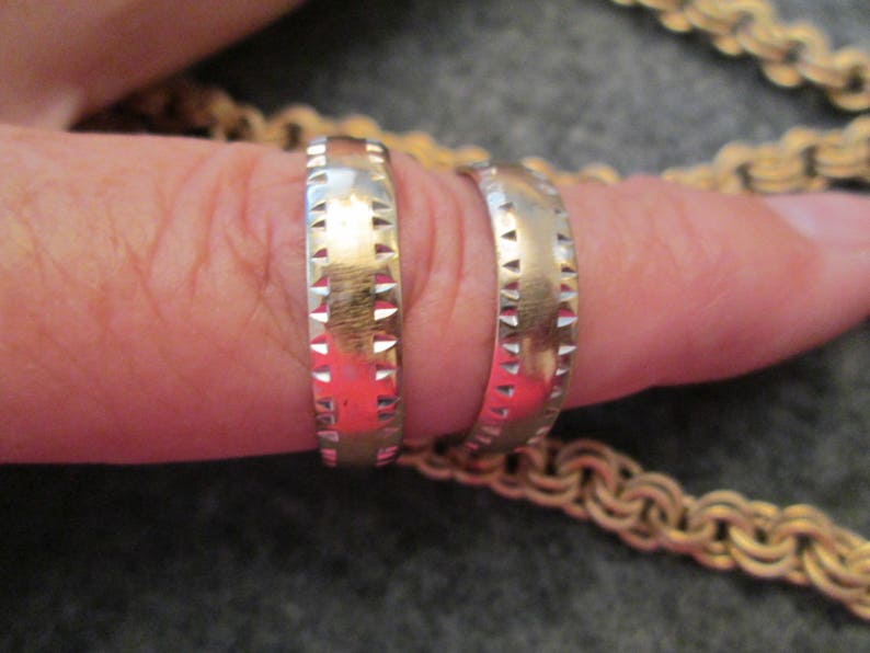 May include: Two gold wedding bands with a textured design. The bands are worn on a finger.