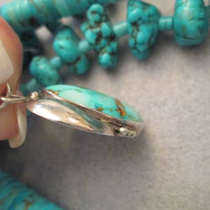 Peut inclure: Un pendentif en turquoise avec un sertissage en argent. Le pendentif est de forme ovale et a une surface lisse et polie. La turquoise est d'une couleur bleu-vert vibrante avec des veines brunes qui la traversent.
