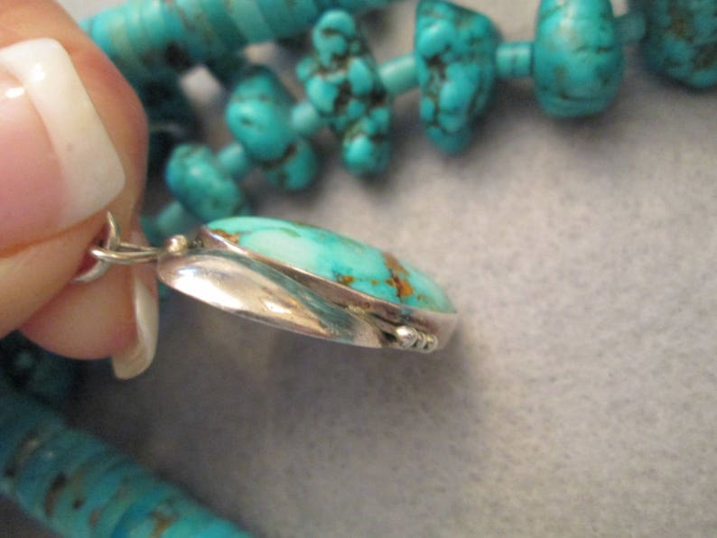 Peut inclure: Un pendentif en turquoise avec un sertissage en argent. Le pendentif est de forme ovale et a une surface lisse et polie. La turquoise est d'une couleur bleu-vert vibrante avec des veines brunes qui la traversent.
