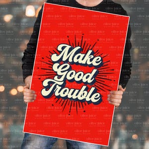 Könnte beinhalten: Ein rotes Poster mit dem Schriftzug "Make Good Trouble" in einer Retro-Schriftart. Der Text ist weiß und marineblau, mit einem Burst-Design im Hintergrund. Das Poster wird von einer Person hochgehalten.