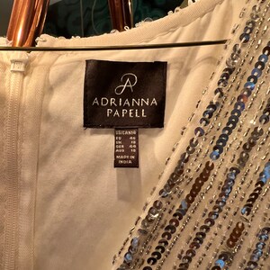 Op de afbeelding: Ivoor kleurige jurk met een kralen- en pailletten ontwerp. Het label luidt "ADRIANNA PAPELL" en bevat maat informatie. De jurk is gemaakt in India.