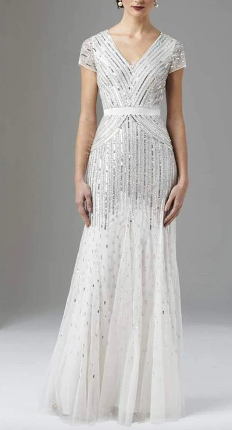 Op de afbeelding: Een witte, vloerlange jurk met een V-hals en korte mouwen. De jurk is rijkelijk versierd met zilveren pailletten en kralen, wat een glinsterend effect cre&euml;ert. Een witte riem accentueert de taille en de rok loopt wijd uit aan de onderkant.