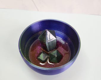 以下が含まれることがあります： 紫色のボウルに、虹色の緑と黒のサイコロが3つ入っています。ボウルは、キラキラ光る濃い紫色のレジンで満たされています。