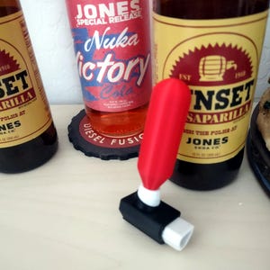 May include: A red and black bottle opener with a white tip, next to bottles of soda. The soda bottles have labels with the text "JONES" and "Nuka Victory Cola". The bottles are brown and orange.