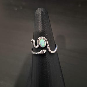 May include: A silver-toned snake ring featuring an oval-shaped iridescent opal stone set in the center. The snake body wraps around the finger with engraved dot details along the band, and the head of the snake is visible on the right side.