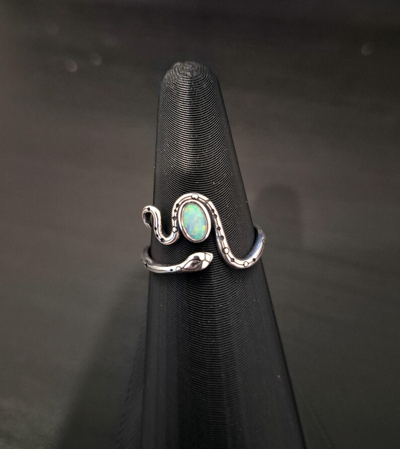 May include: A silver-toned snake ring featuring an oval-shaped iridescent opal stone set in the center. The snake body wraps around the finger with engraved dot details along the band, and the head of the snake is visible on the right side.