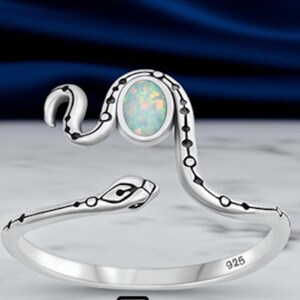 May include: A sterling silver open ring shaped like a coiled snake with a polished finish. The snake features black etched detailing along its body and a central oval-shaped iridescent opal stone. The inner band is clearly stamped with the number 925.