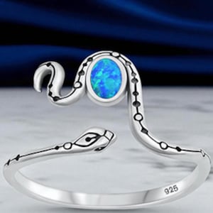 May include: A silver snake-shaped ring featuring a polished finish and a central oval-shaped blue opal stone. The band is engraved with black dotted patterns along the body of the snake, and the interior of the band is stamped with the number 925.