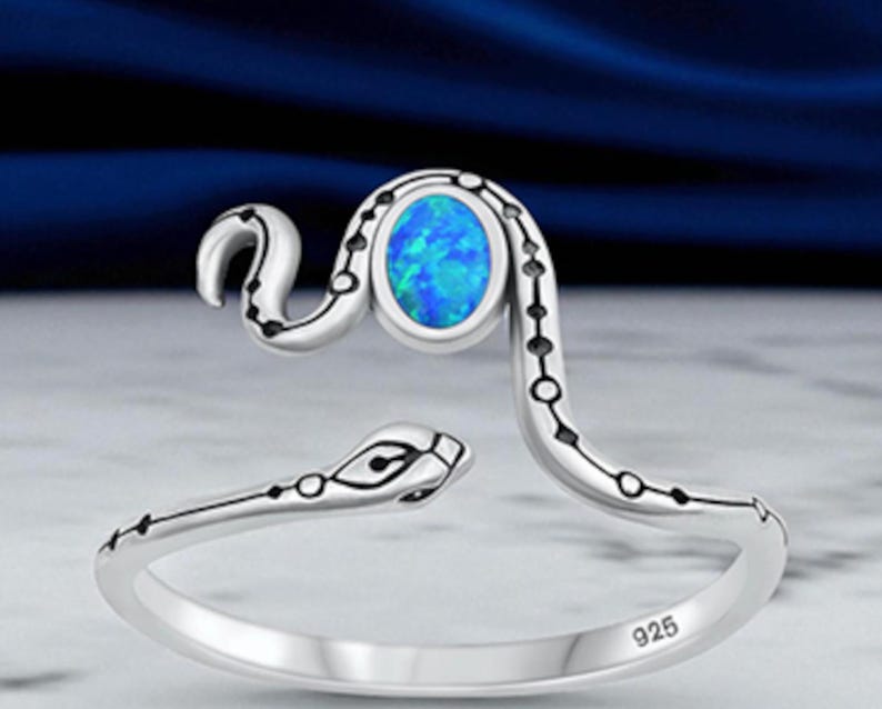 May include: A silver snake-shaped ring featuring a polished finish and a central oval-shaped blue opal stone. The band is engraved with black dotted patterns along the body of the snake, and the interior of the band is stamped with the number 925.