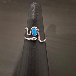 May include: A silver-toned metal ring shaped like a coiled snake with a textured, dotted pattern along its body. The ring features a central, oval-shaped, iridescent blue stone set within the snake's curve, with the head and tail ends overlapping.