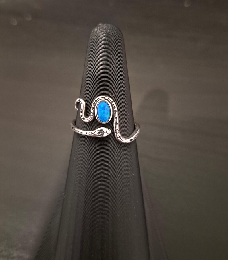May include: A silver-toned metal ring shaped like a coiled snake with a textured, dotted pattern along its body. The ring features a central, oval-shaped, iridescent blue stone set within the snake's curve, with the head and tail ends overlapping.