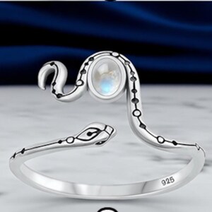 May include: A sterling silver open ring shaped like a snake with a polished finish. The snake's body features small black circular accents along its length, and an oval-shaped iridescent moonstone is set on the head. The band is stamped with 925.