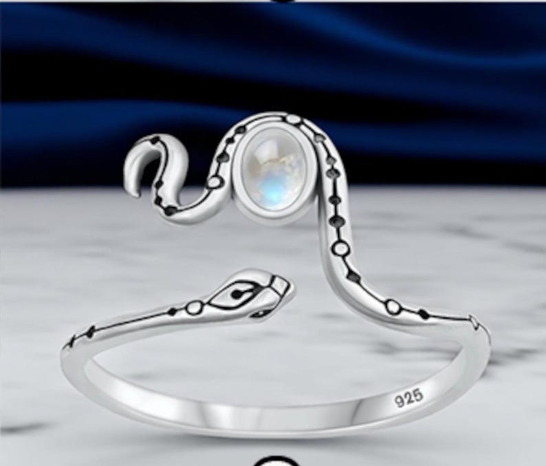 May include: A sterling silver open ring shaped like a snake with a polished finish. The snake's body features small black circular accents along its length, and an oval-shaped iridescent moonstone is set on the head. The band is stamped with 925.