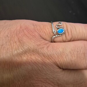 May include: A silver-toned wrap ring shaped like a snake with a textured, oxidized finish. The snake's head is at one end, and the body curves around the finger, featuring a single, oval-shaped, iridescent blue stone set into the center of the band.