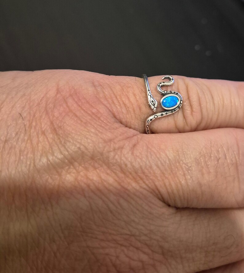 May include: A silver-toned wrap ring shaped like a snake with a textured, oxidized finish. The snake's head is at one end, and the body curves around the finger, featuring a single, oval-shaped, iridescent blue stone set into the center of the band.