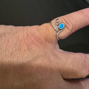 May include: A silver-toned wrap ring shaped like a snake with a textured body and a small, oval-shaped iridescent blue stone set into the head. The metal band curves around the finger, featuring etched details along the serpent's back.