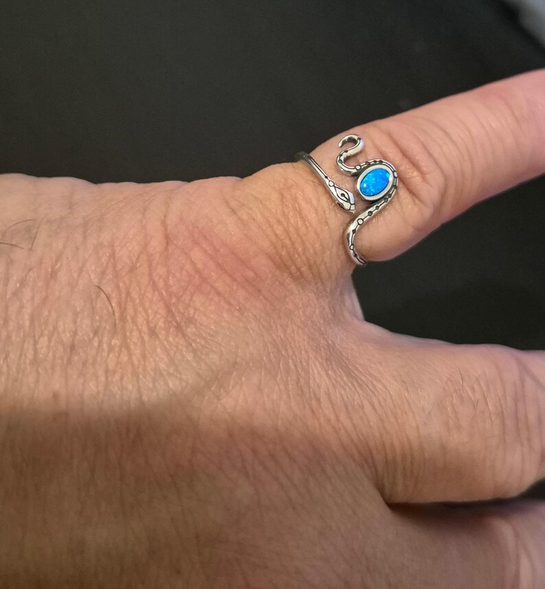 May include: A silver-toned wrap ring shaped like a snake with a textured body and a small, oval-shaped iridescent blue stone set into the head. The metal band curves around the finger, featuring etched details along the serpent's back.