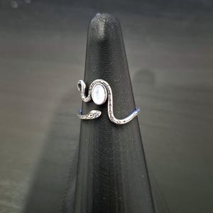 May include: A silver-toned metal ring shaped like a coiled snake with a smooth, oval-shaped white stone set in the center. The band features etched detailing along the body of the snake, including small circular markings and a defined head shape.